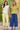 Two women standing side by side in colorful outfits outside, one in yellow-green cropped jeans and a crochet-trimmed top, the other in blue wide leg jeans and a green sweater top.